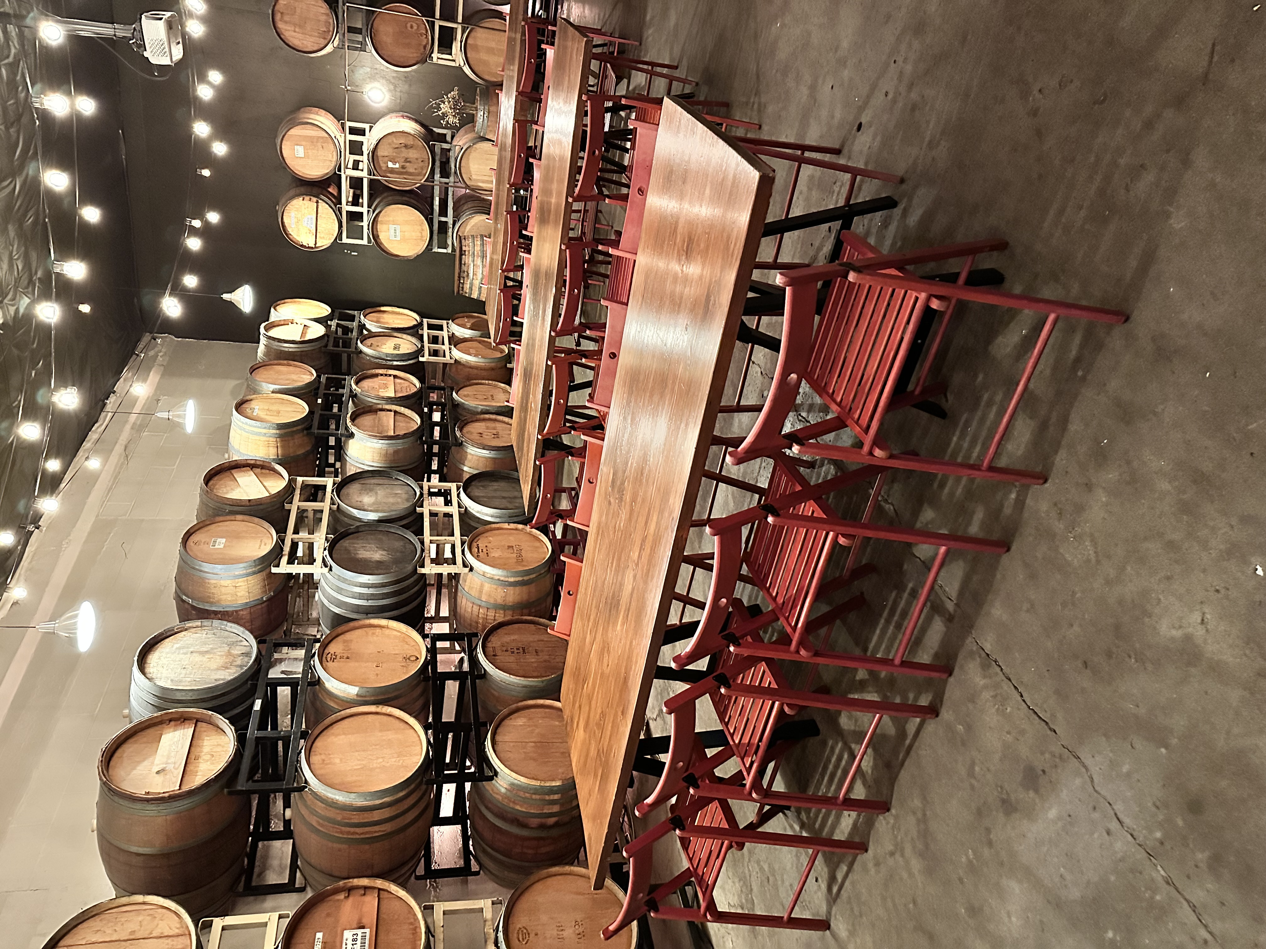 The ENSO Barrel Room set for an event: long farm tables and red folding chairs beneath strings of café lights, surrounded by stacked oak barrels