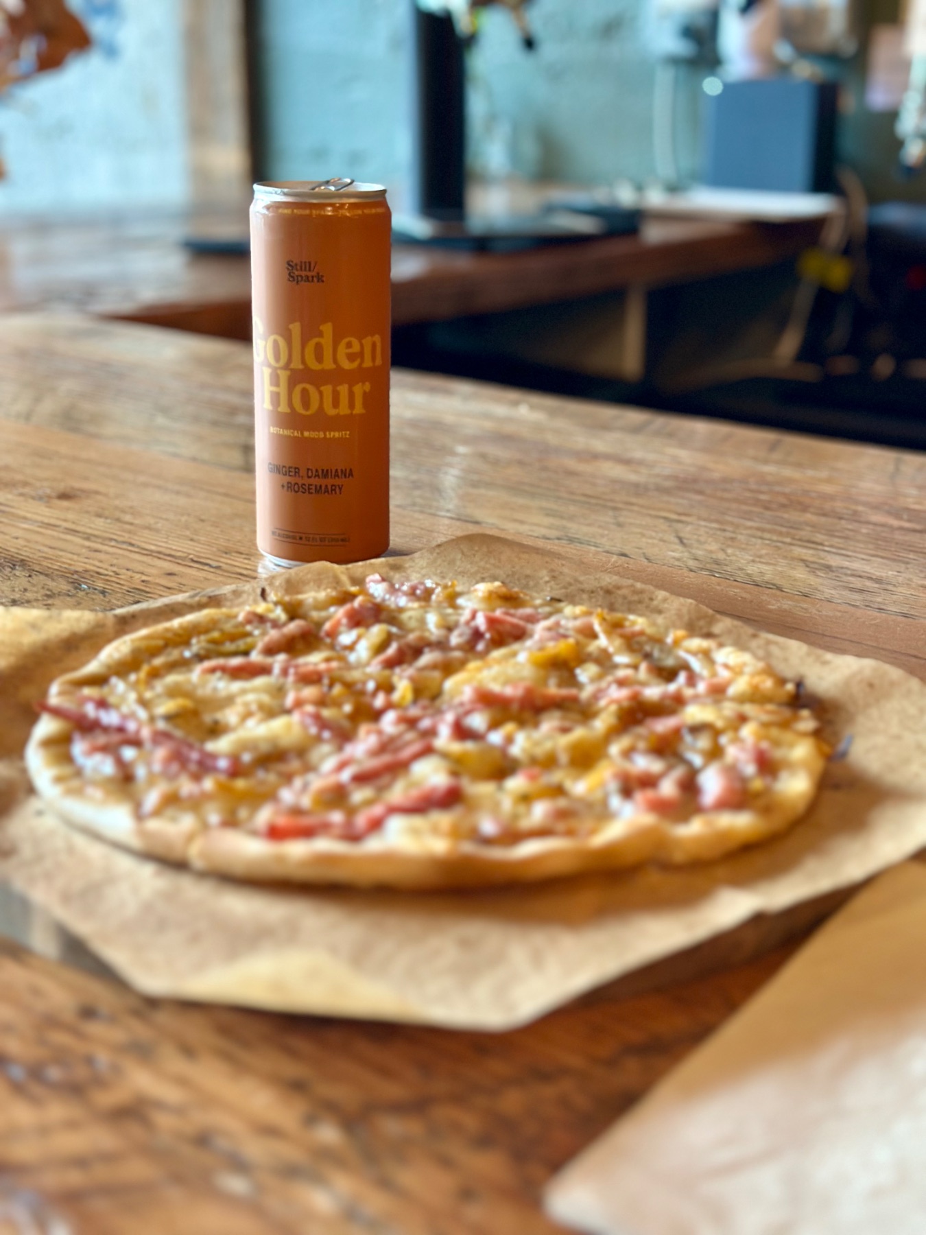 A pizzalette on parchment beside a can of Still/Spark Golden Hour botanical spritz on the ENSO bar