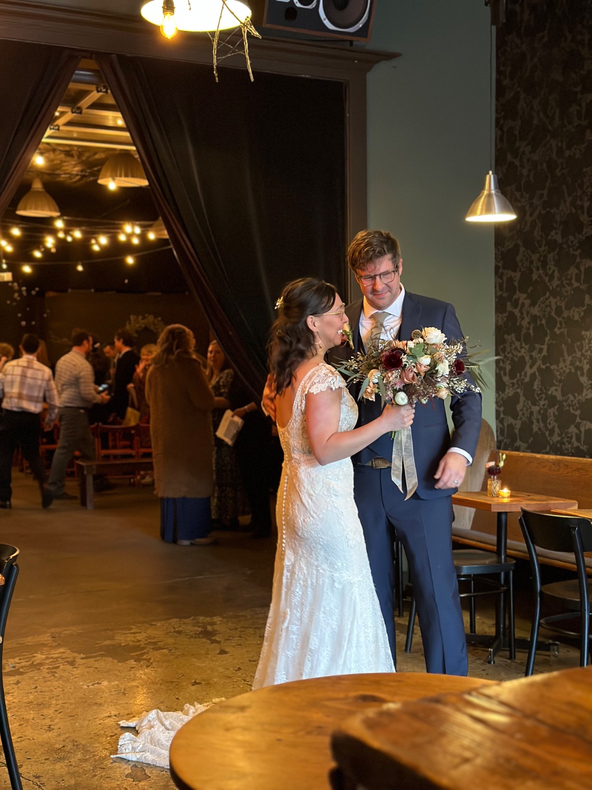 A bride and groom sharing a quiet moment in the Tasting Lounge, her bouquet between them, while guests mingle under the string lights of the Barrel Room behind