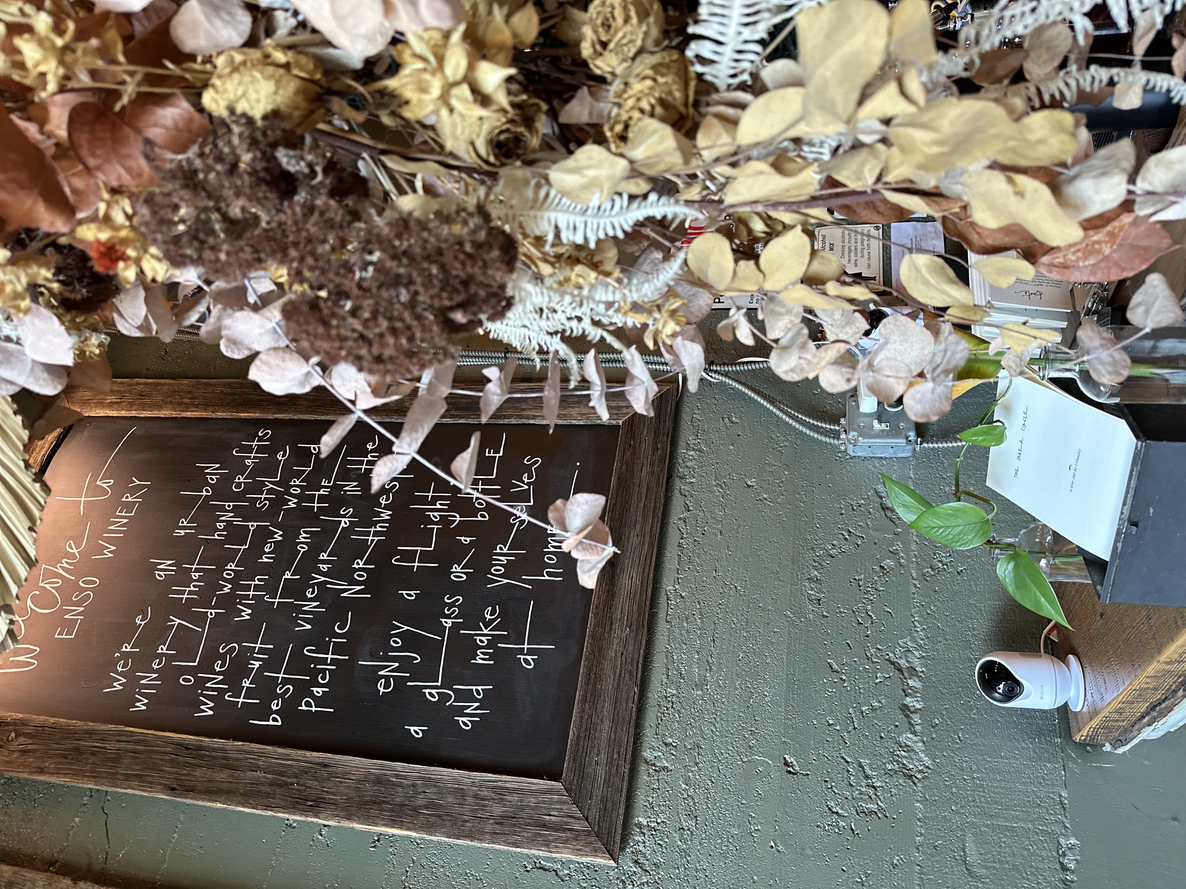 The hand-lettered Welcome to ENSO Winery chalkboard beside a cascade of dried flowers: enjoy a flight, a glass or a bottle, and make yourselves at home